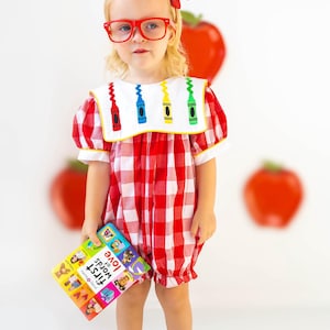 May include: A young child wearing a red and white checkered romper with a white collar featuring crayon designs. The child is wearing red glasses and a red bow. The child is holding a colorful book titled "First Words of Love".