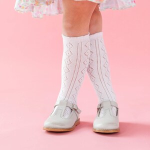 May include: White knee-high socks featuring a vertical diamond-shaped openwork pattern and a ribbed texture. The socks are paired with light gray leather T-strap shoes that have a rounded toe, a single buckle closure, and decorative perforations.