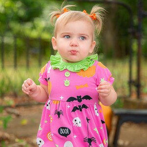 May include: A pink Halloween-themed romper with a green ruffled collar. The romper features a pattern of bats, pumpkins, ghosts, and the word "BOO!". The outfit is worn by a young child with orange hair ties.