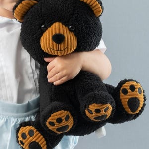 May include: A black plush teddy bear with brown corduroy accents on the ears, nose, and paws. The bear has a friendly expression and is sitting upright.