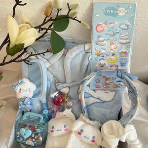 May include: A gift box filled with light blue and white items. Includes a plush toy, a comb, a headband, a scrunchie, a sticker sheet with the text "PINKSODA", a lollipop, and a small container with heart-shaped decorations. A blue robe is also visible.