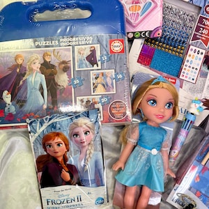 May include: A collection of Frozen II themed toys and accessories. Includes a doll in a blue dress, a puzzle, a beauty kit, and various craft supplies. The items are arranged on a white surface.