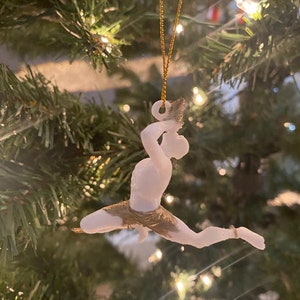 May include: White 3D printed Christmas ornament of a person in a gold-colored shorts, hanging from a gold string on a green Christmas tree.
