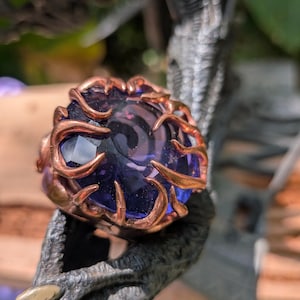 Puede incluir: Un anillo con una gran piedra preciosa facetada de color morado intenso, sostenida por un engaste de metal color cobre que recuerda a ramas curvas o tentáculos en forma de garra que envuelven los bordes de la piedra con un patrón orgánico.