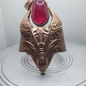 May include: A copper-colored metal ring with an ornate design. The ring features a large, oval, red gemstone at the top. The ring's design includes stylized wings and a detailed face at the base, creating a unique and artistic piece of jewelry.