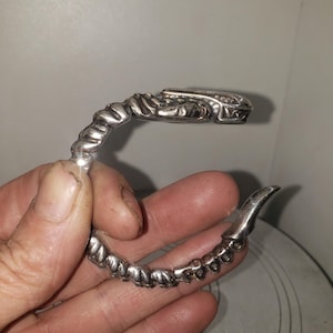 Puede incluir: Un brazalete plateado con un diseño detallado y texturizado. El brazalete se curva en forma de C, con un extremo en forma de garra y una serie de pequeñas formas redondeadas a lo largo de su longitud. El diseño tiene un tema gótico o de fantasía.