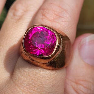 May include: A gold-toned ring featuring a large, oval-cut, vibrant pink gemstone. The ring's band is thick and smooth, with a polished finish. The gemstone is the focal point, catching the light and displaying a rich, saturated color.