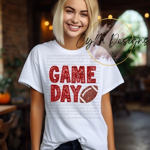 Peut inclure: Un t-shirt blanc avec du texte rouge pailleté qui dit "GAME DAY" et un graphique de ballon de football brun et blanc. Le texte "CYM Designs" est répété plusieurs fois en plus petit autour du graphique.