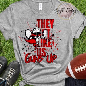 Pode incluir: Uma camiseta cinza com um design gráfico vermelho que apresenta um personagem de desenho animado e o texto "They Not Like Us Guns Up".