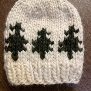 May include: A cream-colored knit beanie with a dark green tree pattern. The hat is made of thick, chunky yarn, and the trees are evenly spaced across the front. The beanie is a winter accessory.