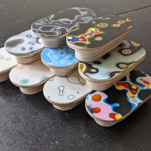 Peut inclure: Une pile de dix sous-verres en bois de forme ovale avec des motifs abstraits colorés. Les sous-verres sont en bois avec un dessus en résine époxy transparente. Les motifs sont tous différents et présentent des volutes, des points et des lignes de différentes couleurs.
