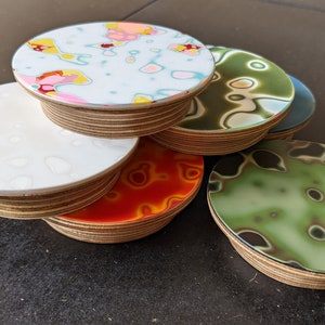 May include: A stack of six round coasters with different abstract designs. The coasters are made of wood with a clear epoxy resin top. The designs include white with gray spots, green with black swirls, orange with red swirls, and blue with white swirls.