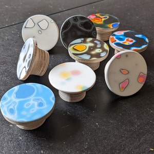May include: A collection of decorative cabinet knobs with colorful, abstract designs. The knobs have a round top with a patterned surface and a wooden base. The color palette includes blue, white, black, yellow, and pink. These knobs are suitable for furniture or home decor.
