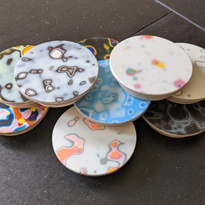 May include: A stack of eight round coasters with abstract patterns in shades of blue, black, white, pink, and orange. The coasters are made of wood and have a glossy finish.