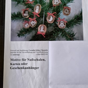 May include: A collection of 10 handmade Christmas ornaments featuring various designs, including a star, a bell, a tree, and a crescent moon. The ornaments are made of wood and decorated with white embroidery thread. They are hanging from a pine branch with red ribbon.