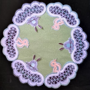Könnte beinhalten: Eine weiße Häkeldeckchen mit grünem Zentrum und gestickten Bildern von Vögeln und Kaninchen.