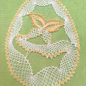 May include: A white lace doily with a yellow border, featuring a delicate floral design. The doily is on a green background.