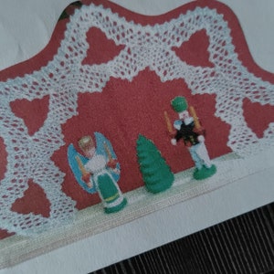 May include: A white crocheted heart-shaped archway with a red background. There are three figurines in the archway: a green Christmas tree, a white angel with a green dress, and a red and white nutcracker.