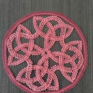 May include: A pink and burgundy crocheted doily with a circular design. The doily has a delicate, intricate pattern of loops and knots.