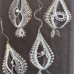May include: Four white lace ornaments with gold accents and a pearl bead in the center of each. The ornaments are hanging from a dark background.