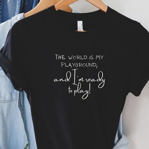 Op de afbeelding: Zwart T-shirt met witte tekst die luidt: "De wereld is mijn speeltuin, en ik ben klaar om te spelen!"