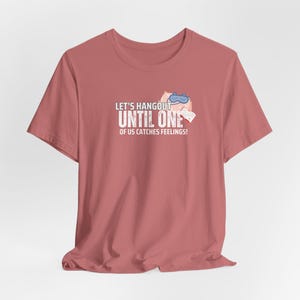May include: A pink t-shirt featuring a graphic of a cat wearing a sleep mask and holding a book. The text on the shirt reads "Let's Hangout Until One Of Us Catches Feelings!"
