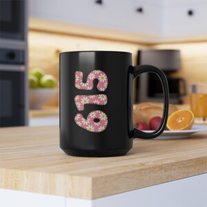 May include: Black ceramic mug with the number "615" in a pink floral pattern. The mug has a large handle and is sitting on a wooden surface. The background includes a kitchen setting with a bowl of fruit.