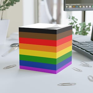 May include: A cube-shaped notepad with a Pride flag design. The notepad features layers of black, brown, red, orange, yellow, green, blue, and purple. A white sheet of paper is on top. Several silver paper clips are scattered around the notepad.