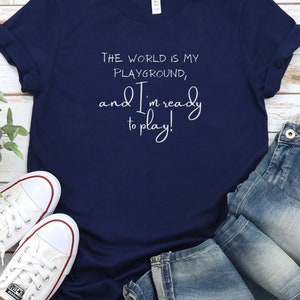 Op de afbeelding: Een marineblauw T-shirt met witte tekst die luidt: "De wereld is mijn speeltuin, en ik ben klaar om te spelen!"