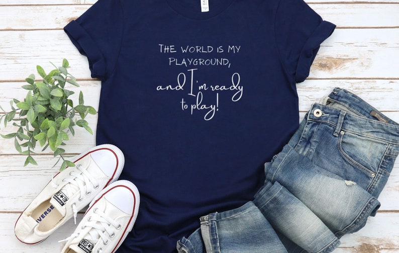 Op de afbeelding: Een marineblauw T-shirt met witte tekst die luidt: "De wereld is mijn speeltuin, en ik ben klaar om te spelen!"