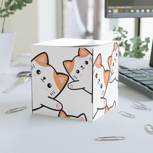 May include: White cube-shaped notepad dispenser with a repeating pattern of cartoon cats in orange and white. The cats have black outlines and smiling faces. The dispenser is on a white desk with paper clips.