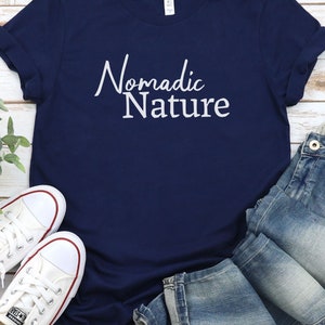 May include: A navy blue t-shirt with the text "Nomadic Nature" in white lettering. The shirt is paired with blue jeans and white sneakers.