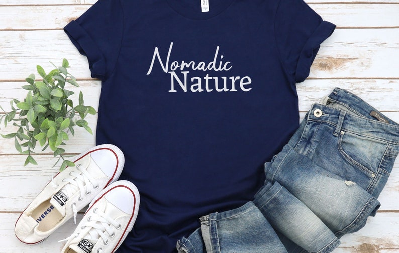 May include: A navy blue t-shirt with the text "Nomadic Nature" in white lettering. The shirt is paired with blue jeans and white sneakers.