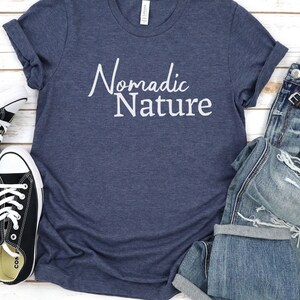 May include: A navy blue t-shirt with the text "Nomadic Nature" in white lettering. The shirt is paired with blue jeans and black sneakers.