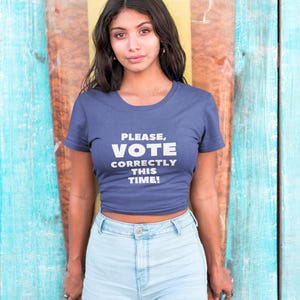 May include: A navy blue crop top with the text "PLEASE, VOTE CORRECTLY THIS TIME!" in white. The top is paired with light blue denim shorts. The background is a colorful wooden surface.