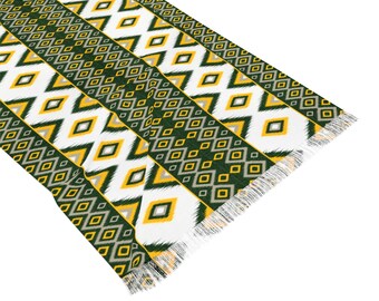 May include: A patterned textile featuring a repeating geometric design in green, yellow, white, and gray. The design includes diamond and zigzag shapes. The textile has a fringed edge, suggesting it could be a scarf, throw, or decorative fabric.