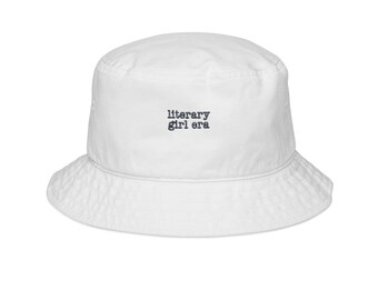 May include: White bucket hat with a rounded crown and a downward-sloping brim. The hat features the text "literary girl era" in a small, centered, black font. The hat is made of a soft, possibly cotton, material.