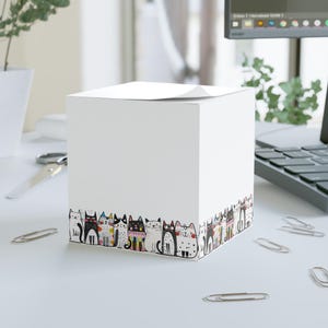 May include: A white cube notepad featuring a border of illustrated cats in various poses and colours. The cats have black, white, and patterned designs. The notepad is on a white desk with office supplies.
