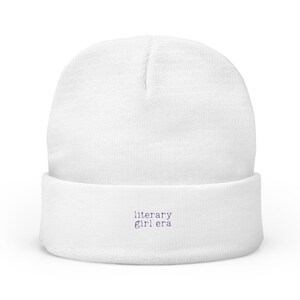 May include: A white beanie hat with a turned-up brim. The words "literary girl era" are embroidered in purple on the brim. The hat is made of a soft, knitted fabric.