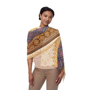 May include: A patterned shawl with a cream-colored base and intricate designs in shades of blue, purple, gold, and brown. The shawl features geometric and floral patterns, with a fringed edge. The woman is wearing a beige top and brown pants.
