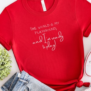 Op de afbeelding: Rood T-shirt met korte mouwen met witte tekst die luidt: "De wereld is mijn speeltuin, en ik ben klaar om te spelen!"