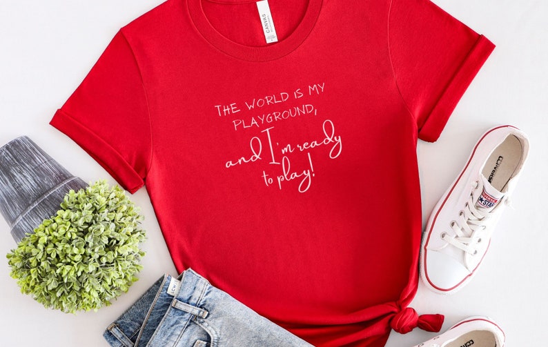 Op de afbeelding: Rood T-shirt met korte mouwen met witte tekst die luidt: "De wereld is mijn speeltuin, en ik ben klaar om te spelen!"
