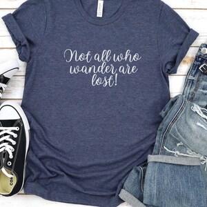 May include: A navy blue t-shirt with the text "Not all who wander are lost!" printed in white on the front. The shirt is paired with distressed denim jeans and black canvas sneakers.