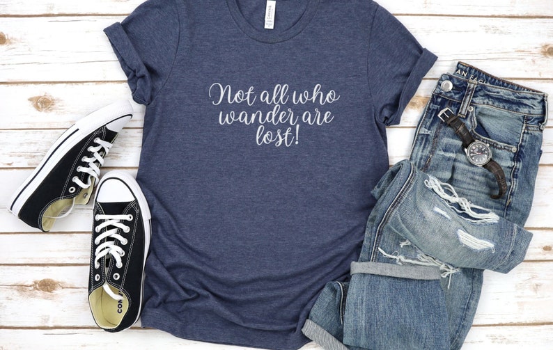 May include: A navy blue t-shirt with the text "Not all who wander are lost!" printed in white on the front. The shirt is paired with distressed denim jeans and black canvas sneakers.