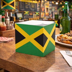 May include: A square notepad holder with the Jamaican flag design. The holder is green with a yellow cross and black diamond shapes. White paper is visible on top. The holder sits on a wooden table.