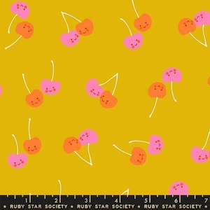 May include: A mustard yellow fabric featuring a repeating pattern of smiling pink and orange cherries with white stems. A black ruler at the bottom edge displays measurements in inches, with the text Ruby Star Society printed repeatedly.