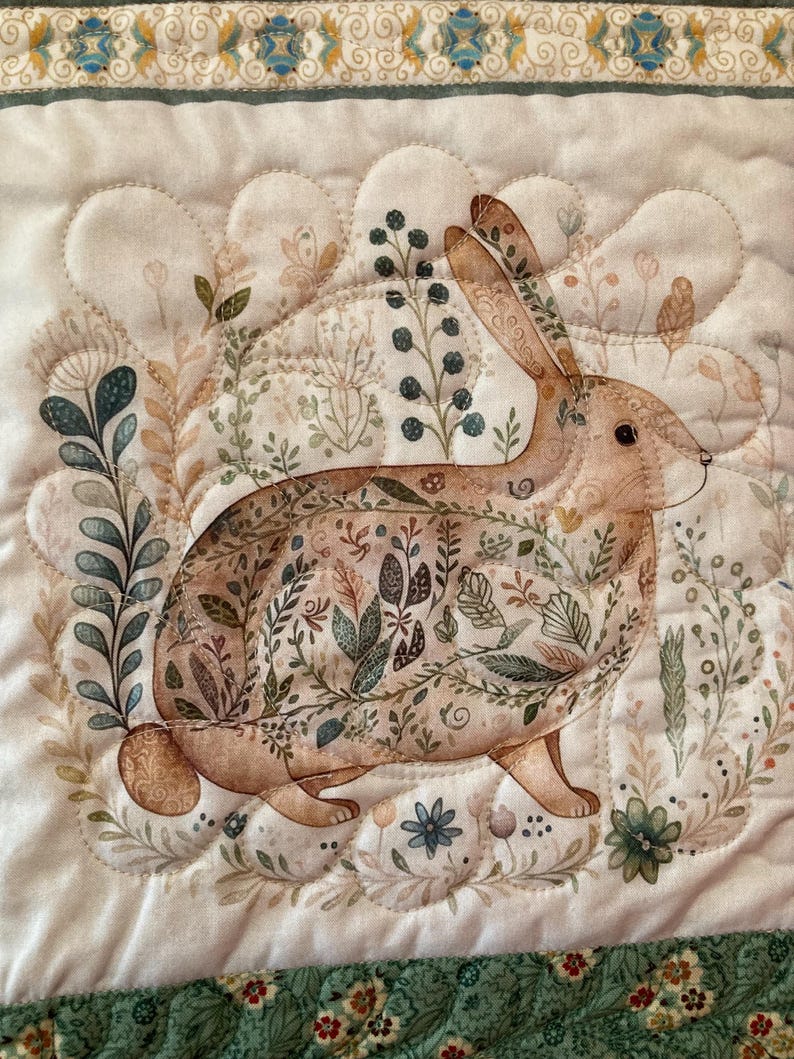 以下が含まれることがあります： 茶色のウサギと花柄のキルト生地。ウサギは、緑、茶色、ベージュの色合いの植物イラストに囲まれています。キルトはスカラップエッジと、花柄とスクロールデザインの装飾的なボーダーが付いています。