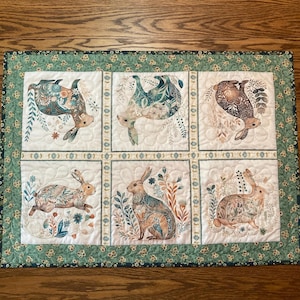 以下が含まれることがあります： 6つのパネルにウサギのイラストが描かれたキルトのテーブルランナー。各ウサギは、青、茶色、ベージュの色合いの花柄で装飾されています。ランナーは、花柄のデザインの緑色のボーダーが付いています。