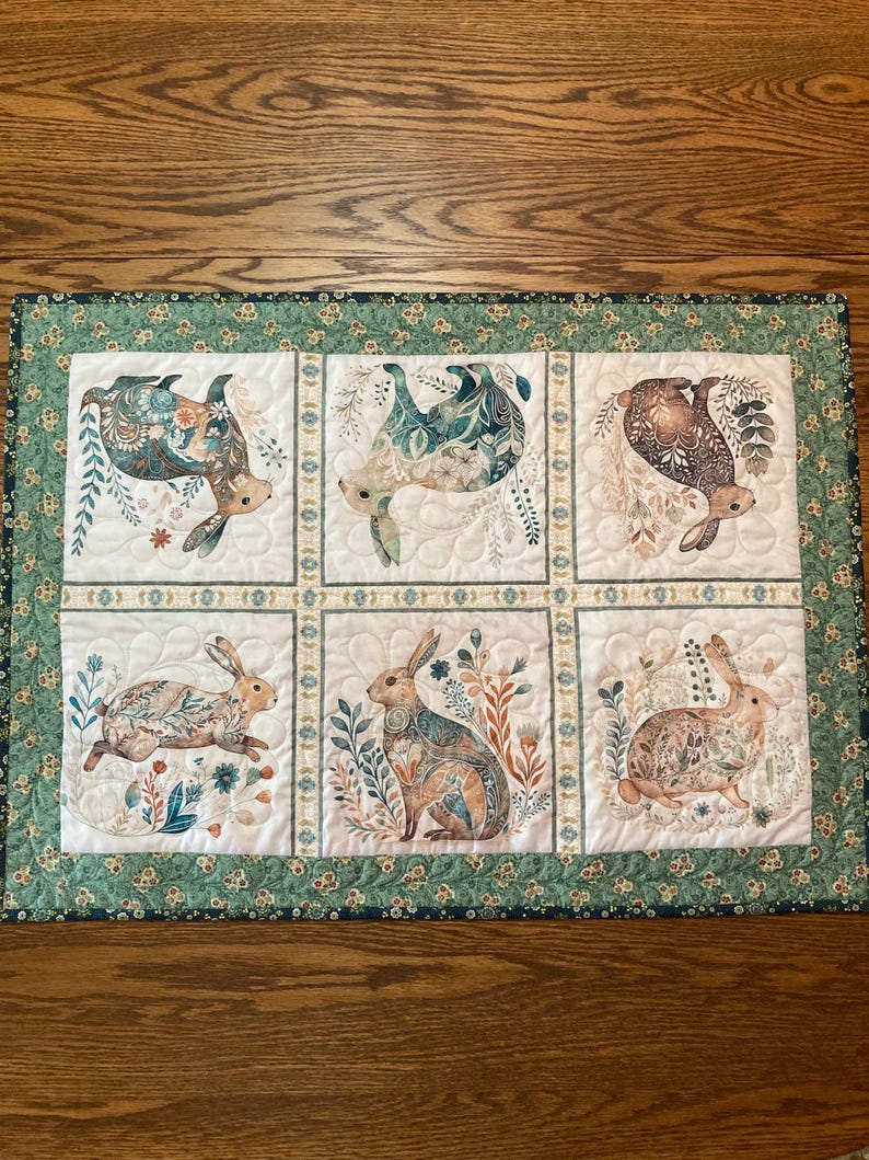 以下が含まれることがあります： 6つのパネルにウサギのイラストが描かれたキルトのテーブルランナー。各ウサギは、青、茶色、ベージュの色合いの花柄で装飾されています。ランナーは、花柄のデザインの緑色のボーダーが付いています。