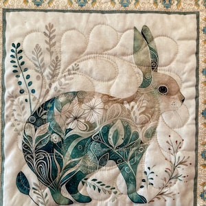 以下が含まれることがあります： ウサギの詳細なイラストが描かれた、キルト加工された布の正方形。ウサギは、ティールと白の花柄で埋められています。背景は白で、ティールと金のフローラルデザインの装飾的なボーダーが付いています。キルトはティール、茶色、白のミックスです。